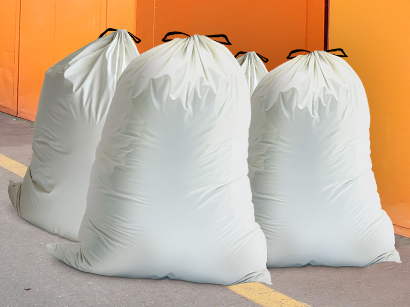Vier volle Kehrichtsäcke stehen auf dem Boden vor einem orangen Container – steht für regulären Hausmüll, der nicht in Recyclingcontainer gehört und über die offizielle Abfallsammlung entsorgt werden muss