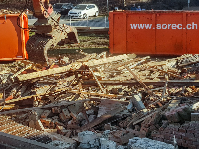 Bagger sortiert gemischten Bauschutt vor orangefarbenem SoRec-Container – zeigt die fachgerechte Entsorgung und Trennung von Bauabfällen auf einer Rückbau- oder Sanierungsbaustelle