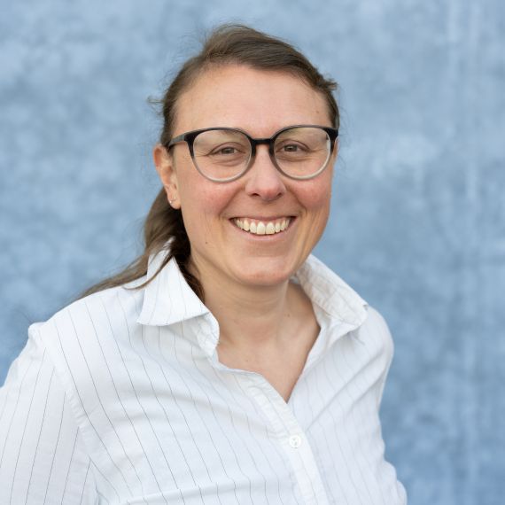 Das Bild zeigt eine lächelnde Frau mit Brille und zusammengebundenen, braunen Haaren, die ein weisses Hemd mit feinem Muster trägt. Sie steht vor einem hellblauen Hintergrund mit leichter Textur. Sie arbeitet in der Administration der soRec AG und heisst Melanie Schwizer.